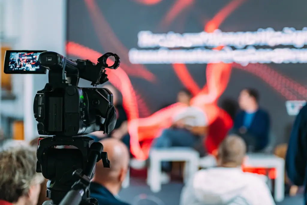 filming-panel-discussion-with-camera-in-foreground-2026-03-25-10-21-40-utc-microgen-via-envato professionelle Kamera, die eine unscharf fotografirete Veranstaltung filmt als Symbolbild für Hybrie Videokonferenzen